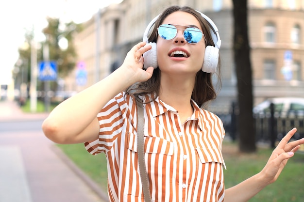 Sonriente mujer hermosa joven feliz en auriculares caminando al aire libre en la ciudad.