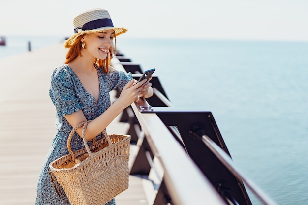 Sonriente mujer hermosa permanecer en el muelle y navegar por teléfono inteligente en un día soleado con ropa de moda ...