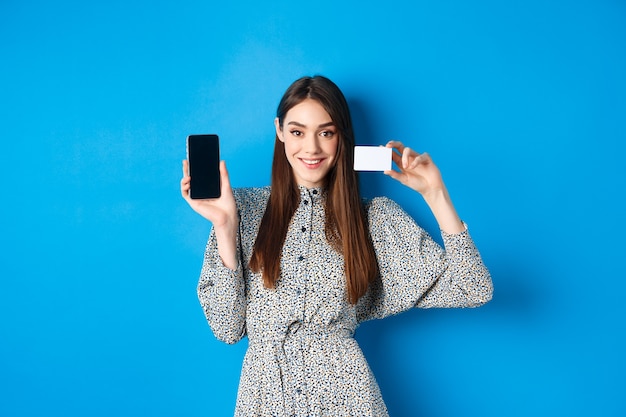 Sonriente mujer hermosa en vestido mostrando teléfono inteligente y tarjeta de crédito de plástico