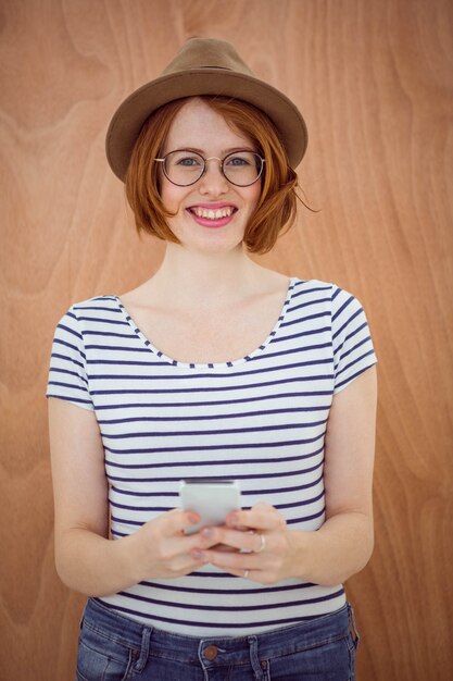 sonriente mujer hipster sosteniendo un teléfono móvil y de pie delante de una madera