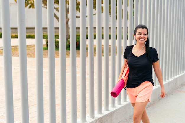 Sonriente mujer hispana escuchando música mientras lleva una estera de yoga