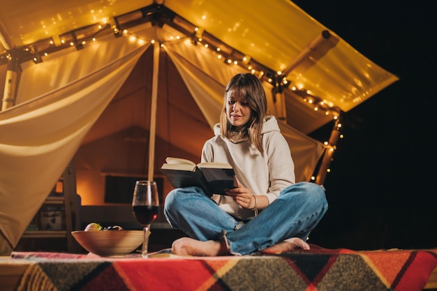 Sonriente mujer independiente bebiendo vino y leyendo un libro sentado en una acogedora carpa glamping en la noche de otoño Tienda de campaña de lujo para vacaciones al aire libre y vacaciones Concepto de estilo de vida