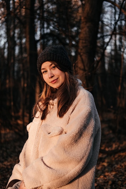 Sonriente a mujer joven en abrigo y sombrero de pie en el bosque de otoño