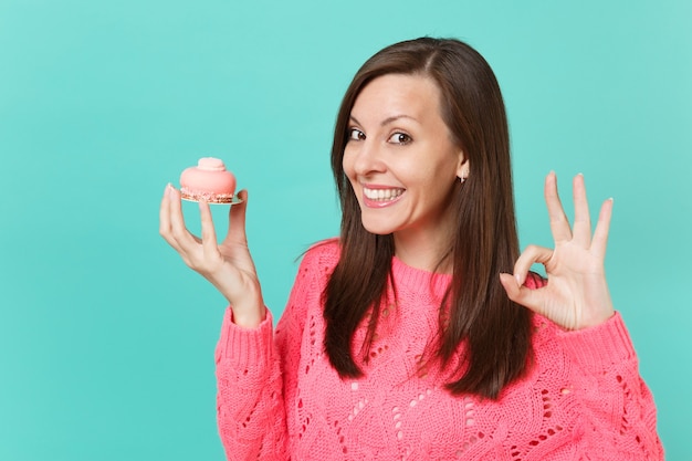 Sonriente a mujer joven atractiva en suéter rosa de punto que muestra el gesto de OK sosteniendo en la mano pastel aislado en el retrato de estudio de fondo de pared azul turquesa. Concepto de estilo de vida de personas. Simulacros de espacio de copia.