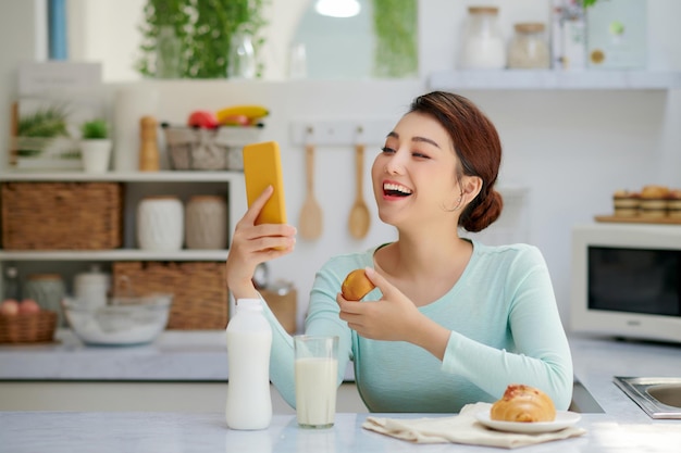 Sonriente mujer joven bastante asiática navegando en Internet mientras desayuna saludablemente en casa