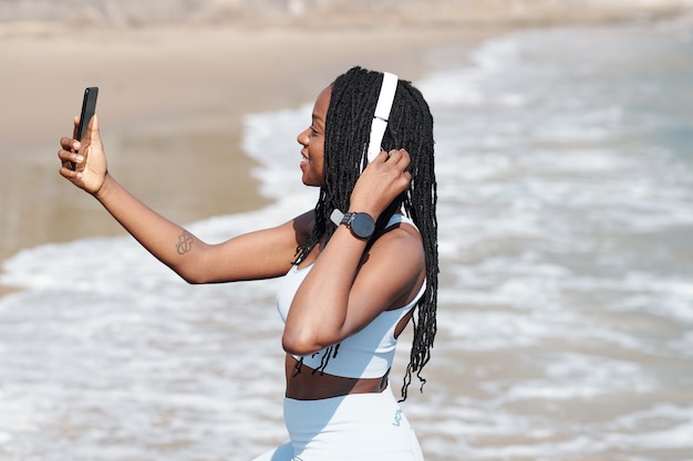 Sonriente mujer joven bastante en forma tomando selfie en la playa después de trotar por la mañana