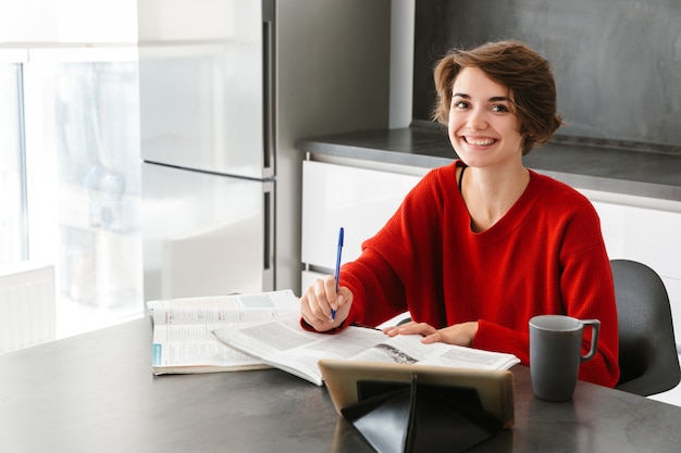 Sonriente a mujer joven y bonita que estudia con una tableta en la mesa en una cocina en casa