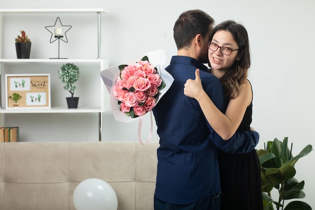 Sonriente a mujer joven y bonita en vasos sosteniendo un ramo de flores y pulgar hacia arriba abrazando a un hombre guapo de pie en la sala de estar