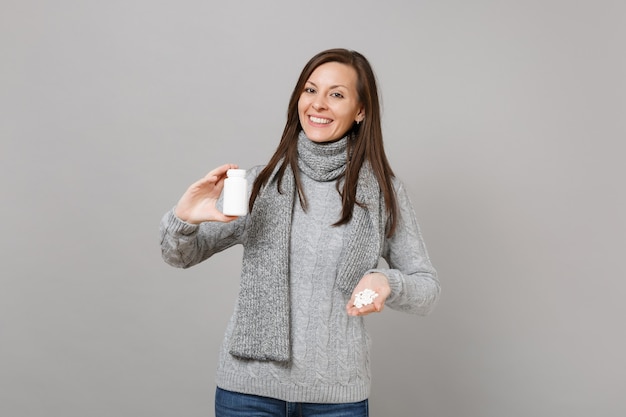 Sonriente a mujer joven en bufanda de suéter gris con tabletas de medicación, píldoras de aspirina en botella aislada sobre fondo gris en estudio. Estilo de vida saludable, tratamiento de enfermedades enfermas, concepto de estación fría.