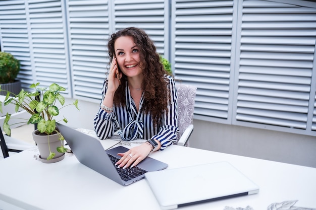 Sonriente a mujer joven exitosa hablando por teléfono, trabajando en una computadora portátil en la terraza, chica en un café en una reunión de negocios. Concepto de negocio