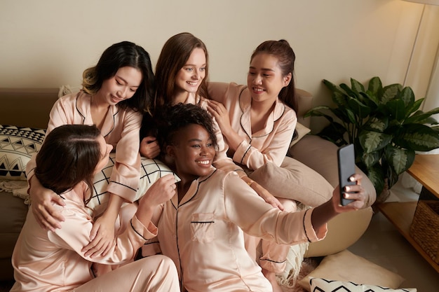 Sonriente mujer joven feliz en pijama de seda tomando selfie con amigos alrededor