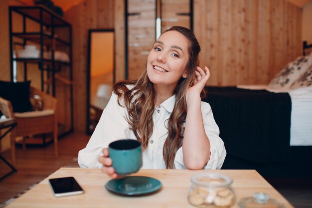 Sonriente mujer joven feliz sentada en la mesa en casa detrás de la computadora portátil y hablando por videollamada chica mujer con taza de té o café hablando en línea en la cámara web en el interior