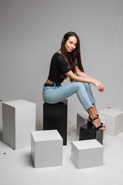 Sonriente a mujer joven en forma con cabello largo posando sentada en cubos blancos y negros sobre fondo gris de estudio con espacio de copia