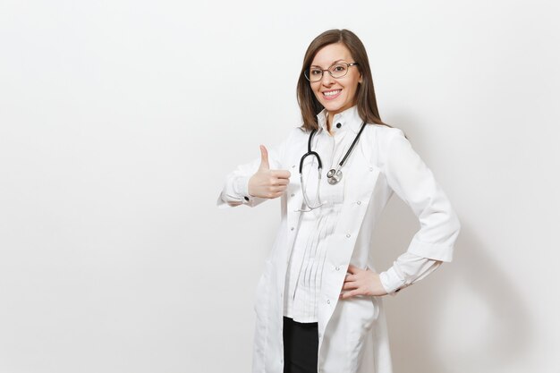Sonriente mujer joven hermosa experimentada médico con estetoscopio, gafas aisladas sobre fondo blanco. Doctora en bata médica muestra los pulgares para arriba. El personal sanitario, la salud, el concepto de medicina.