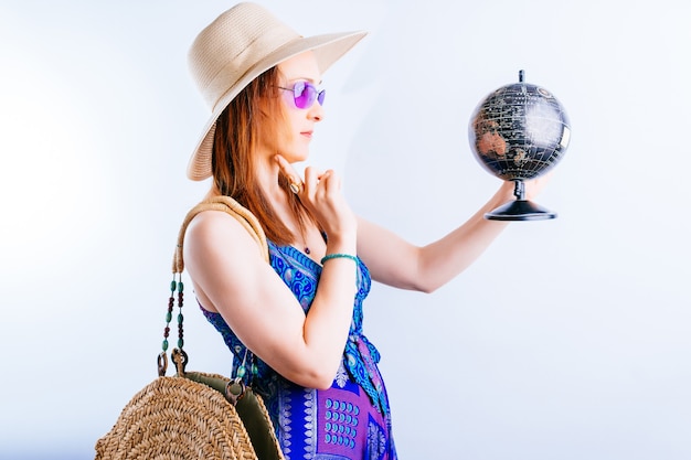 Sonriente a mujer joven hermosa en vestido de playa de verano sosteniendo un globo pensativo viendo dónde viajar en verano. concepto de destino de viaje sobre fondo blanco