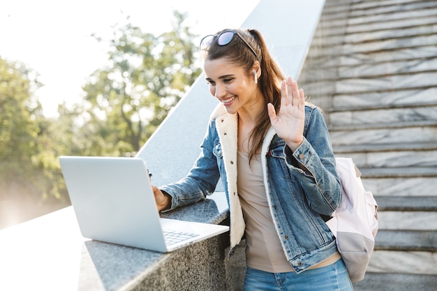 Sonriente, mujer joven, llevando, chaqueta, posición, en, escaleras, aire libre, utilizar, computadora de computadora portátil, ondulación