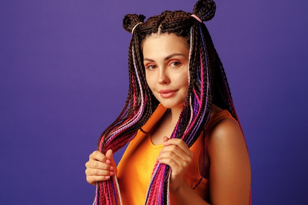 Sonriente a mujer joven positiva con coloridas trenzas africanas sobre fondo púrpura