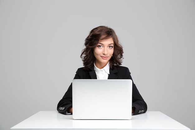 Sonriente a mujer joven rizada hermosa en chaqueta negra sentado y usando la computadora portátil