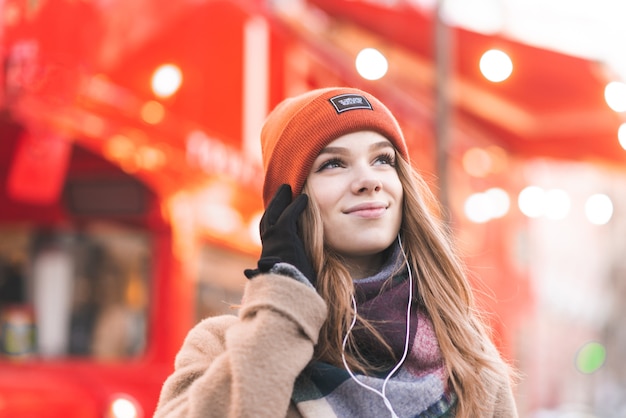 Sonriente mujer joven en ropa de abrigo y un teléfono inteligente en sus manos escucha música en los auriculares y mira hacia los lados en el fondo de un autobús rojo turístico