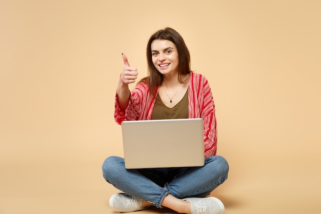 Sonriente a mujer joven en ropa casual mostrando el pulgar hacia arriba, sentado, usando la computadora portátil aislada sobre fondo beige pastel en estudio. Personas sinceras emociones, concepto de estilo de vida. Simulacros de espacio de copia.