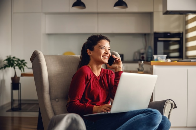 Foto sonriente a mujer joven sentada en casa y tener conversación telefónica con su jefe. en el regazo sostiene la computadora portátil. negocio remoto durante el concepto de virus corona.