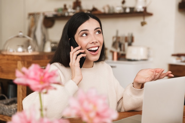 Sonriente a mujer joven sentada en la cocina y usando una computadora portátil, hablando por teléfono móvil