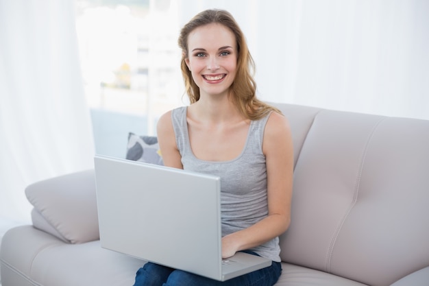 Sonriente mujer joven sentada en el sofá usando la computadora portátil