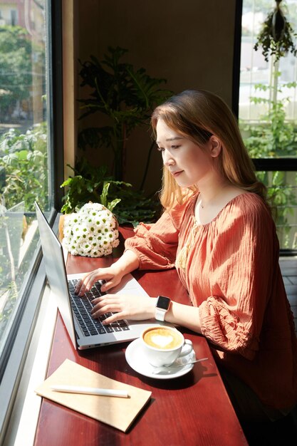 Sonriente a mujer joven sentada en la ventana de la cafetería y los rayos del sol y trabajando en la computadora portátil