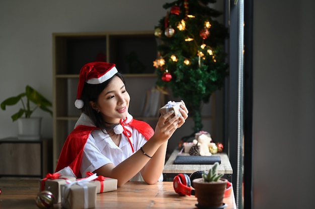 Sonriente a mujer joven con sombrero de Santa con caja de regalo de Navidad mientras está sentado cerca del árbol de Navidad en casa.