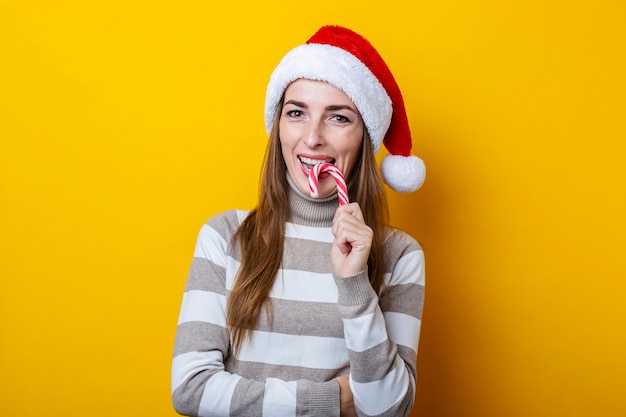 Sonriente a mujer joven con sombrero de Santa Claus comiendo dulces de Navidad sobre fondo amarillo.