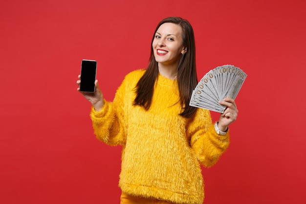 Sonriente a mujer joven sosteniendo teléfono móvil con ventilador de pantalla vacía en blanco de dinero en billetes de dólar dinero en efectivo aislado sobre fondo rojo. Personas sinceras emociones, concepto de estilo de vida. Simulacros de espacio de copia.