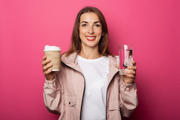 Sonriente a mujer joven sosteniendo un vaso de papel y un vaso con agua sobre la superficie rosa