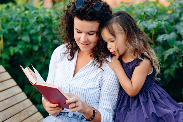 Sonriente mujer joven y su hija leyendo un libro sentado en un banco del parque