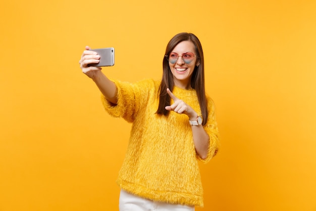 Sonriente a mujer joven en suéter, gafas de corazón haciendo tomar selfie disparo en teléfono móvil apuntando con el dedo índice aislado sobre fondo amarillo brillante. Personas sinceras emociones, estilo de vida. Área de publicidad.