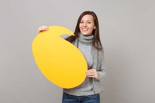 Sonriente a mujer joven en suéter gris, bufanda con amarillo en blanco vacío Diga el bocadillo de diálogo de nube aislado sobre fondo de pared gris. Estilo de vida de moda saludable, personas emociones sinceras, concepto de estación fría.