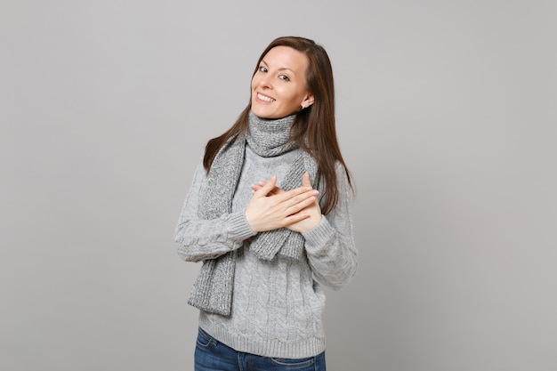 Sonriente a mujer joven en suéter gris, bufanda cogidos de la mano doblados sobre el corazón aislado sobre fondo gris de la pared. Estilo de vida de moda saludable, personas emociones sinceras, concepto de estación fría. Simulacros de espacio de copia.