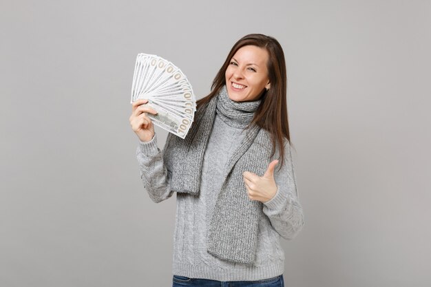 Sonriente a mujer joven en suéter gris, bufanda mostrando el pulgar hacia arriba mantenga un montón de dinero en efectivo de billetes de dólares aislado sobre fondo gris. Emociones de personas de estilo de vida de moda saludable, concepto de estación fría.