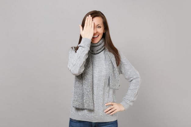 Sonriente a mujer joven en suéter gris, bufanda que cubre los ojos con la mano aislada sobre fondo de pared gris en estudio. Gente de estilo de vida de moda saludable emociones sinceras concepto de estación fría. Simulacros de espacio de copia.