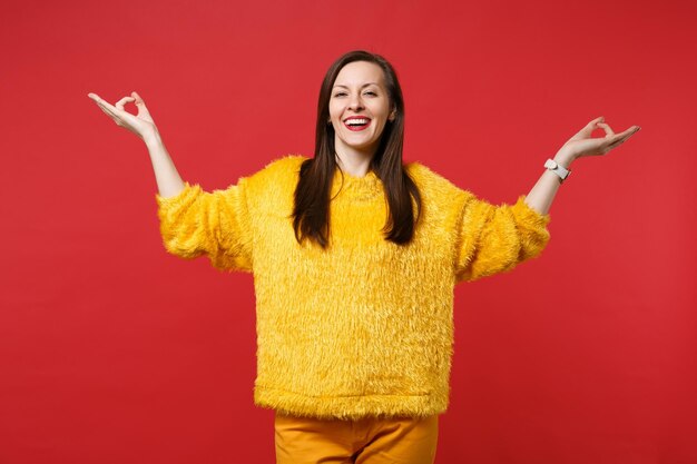 Sonriente a mujer joven en suéter de piel amarilla cogidos de la mano en gesto de yoga, relajante meditando aislado sobre fondo de pared roja en estudio. Personas sinceras emociones, concepto de estilo de vida. Simulacros de espacio de copia.