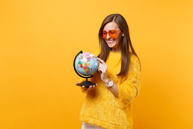 Sonriente a mujer joven en suéter de piel y anteojos de corazón naranja manteniendo el dedo índice en el globo terráqueo aislado sobre fondo amarillo brillante. Personas sinceras emociones, concepto de estilo de vida. Área de publicidad.