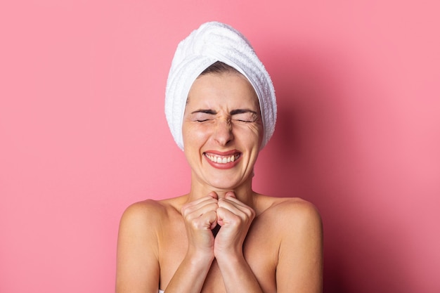 Sonriente a mujer joven con una toalla en la cabeza y los ojos firmemente sobre fondo rosa