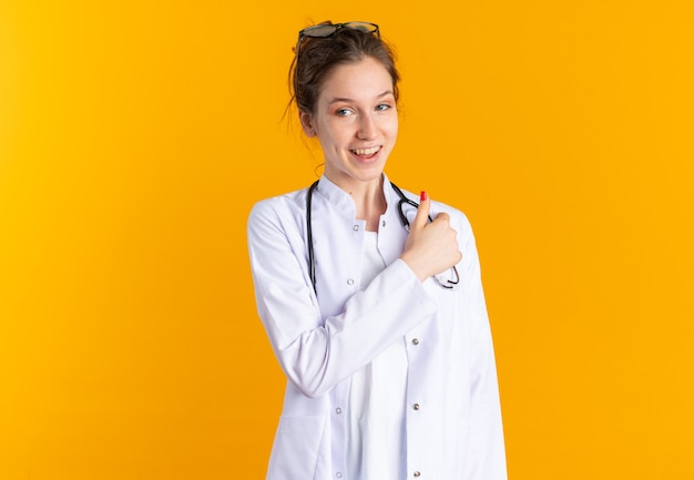 Sonriente a mujer joven en uniforme médico con estetoscopio apuntando a su hombro y mirando al lado