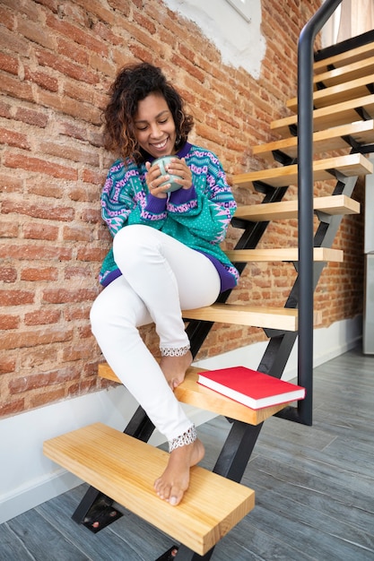 Sonriente mujer latina sosteniendo una taza de café en sus manos. Ella está sentada en las escaleras de su casa junto a un libro.