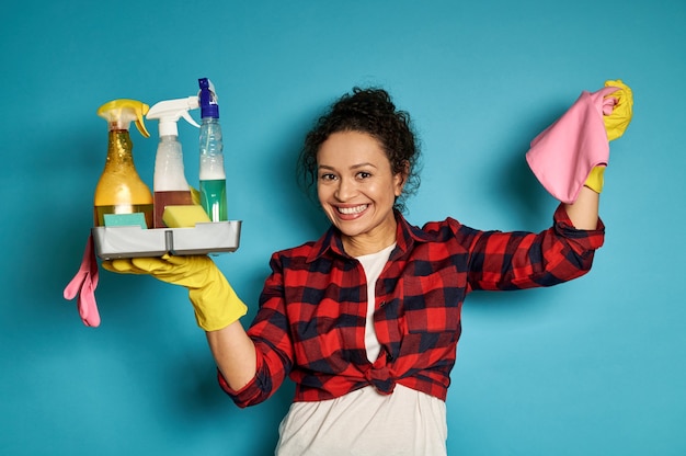 Sonriente mujer latinoamericana levantando los brazos con un trapo rosa y una bandeja llena de productos de limpieza químicos