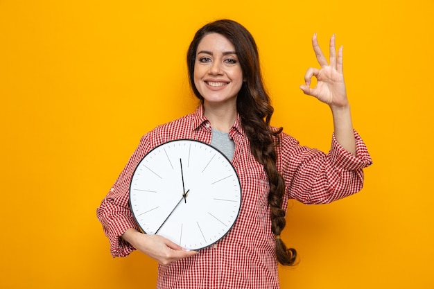 Sonriente mujer limpiador bastante caucásica sosteniendo el reloj y gesticulando el signo de ok con los dedos