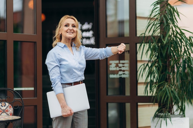 Sonriente mujer madura empresaria sosteniendo portátil contra caffe Mujer exitosa Concepto de trabajador de oficina