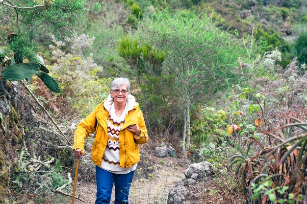 Sonriente mujer mayor al aire libre en excursión a la montaña vistiendo suéter de invierno y chaqueta amarilla. Anciana relajada recostada en bastón disfrutando de la libertad y la naturaleza