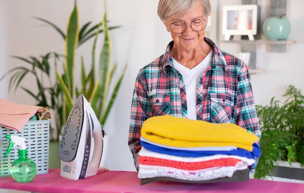 Sonriente mujer mayor atractiva en camisa a cuadros y gafas planchando ropa en casa en tabla de planchar