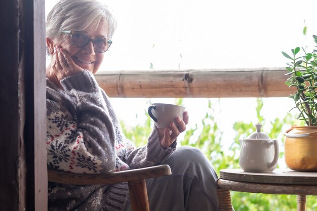 Foto sonriente mujer mayor de pelo gris con un suéter de invierno sentado en el balcón de madera mirando a la cámara sosteniendo una taza de té.