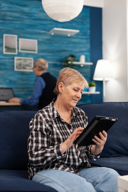 Sonriente mujer mayor con tablet pc descansando en el sofá anciana con tecnología moderna tablet pc en ...